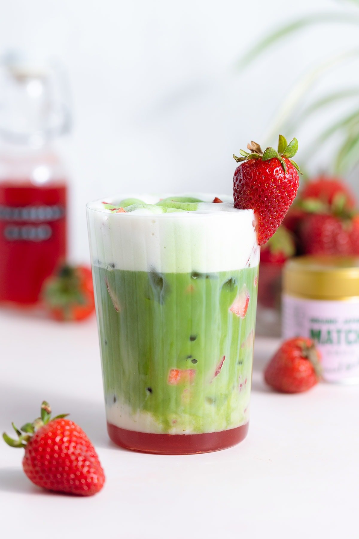 A tall glass with a matcha latte with a layer of frothy milk on the top and a layer of strawberry syrup on the bottom of the glass.