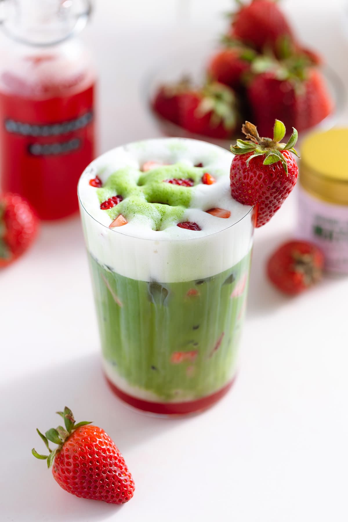 A tall glass with a matcha latte with a layer of frothy milk on the top and a layer of strawberry syrup on the bottom of the glass garnished with more chopped strawberries on top.