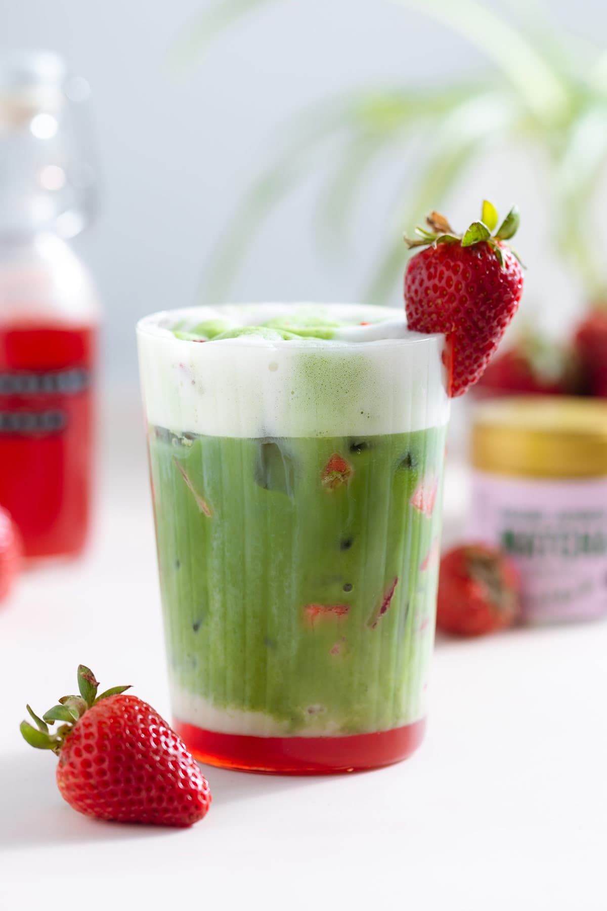 A tall glass with a matcha latte with a layer of frothy milk on the top and a layer of strawberry syrup on the bottom of the glass, garnished with a strawberry.