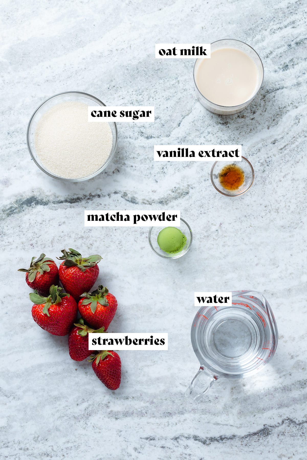 Ingredients like strawberries, milk, cane sugar, and matcha laid out on a grey stone background.