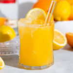 A short glass with bright orange citrus cocktail on a grey background with more lemons and oranges in the background.