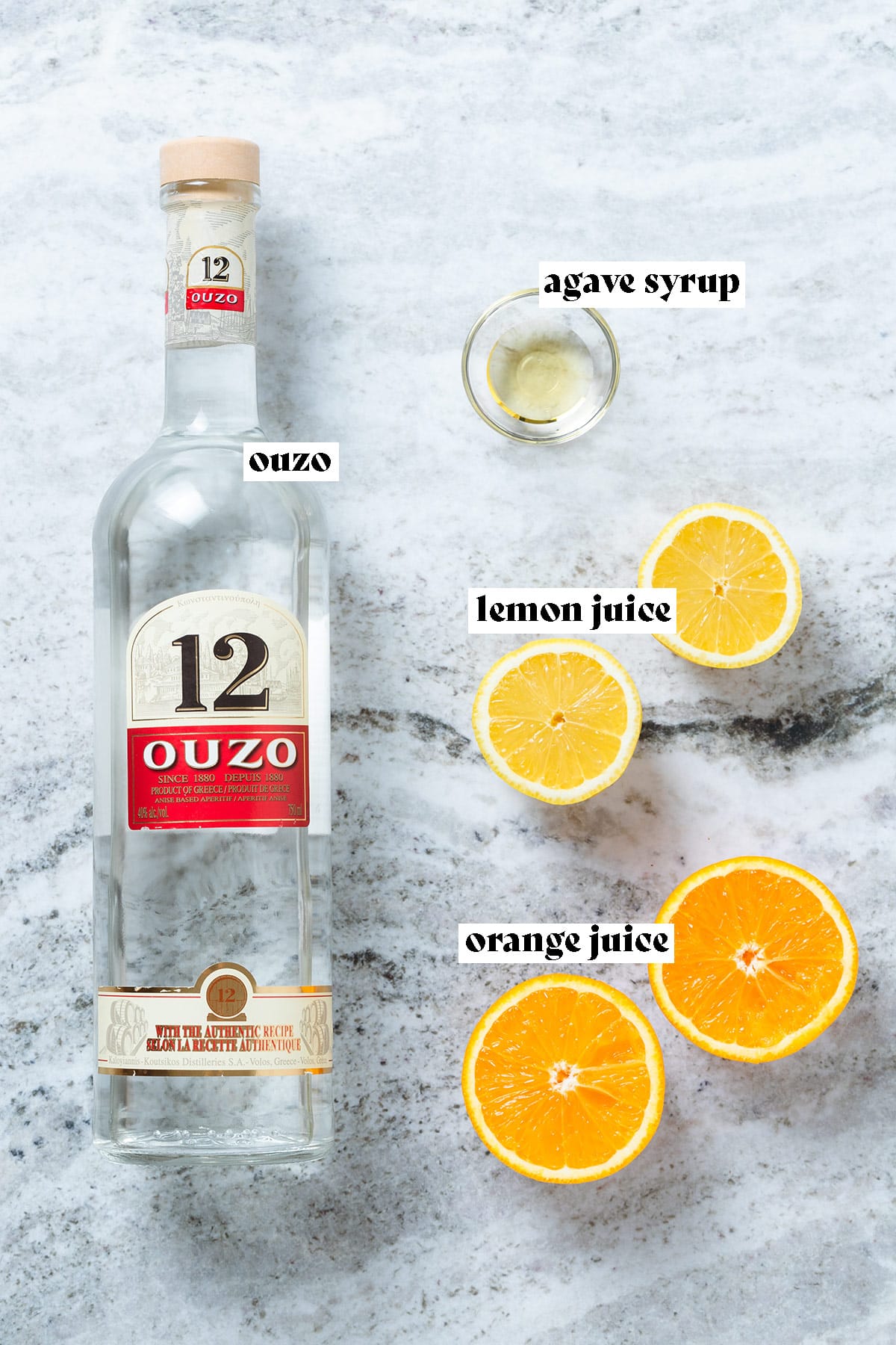 A bottle of Ouzo, halved lemon, orange, and agave syrup on a grey stone background.