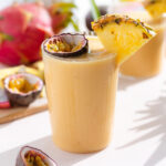 Light orange smoothie in a tall glass garnished with a slice of pineapple and half of a passion fruit.