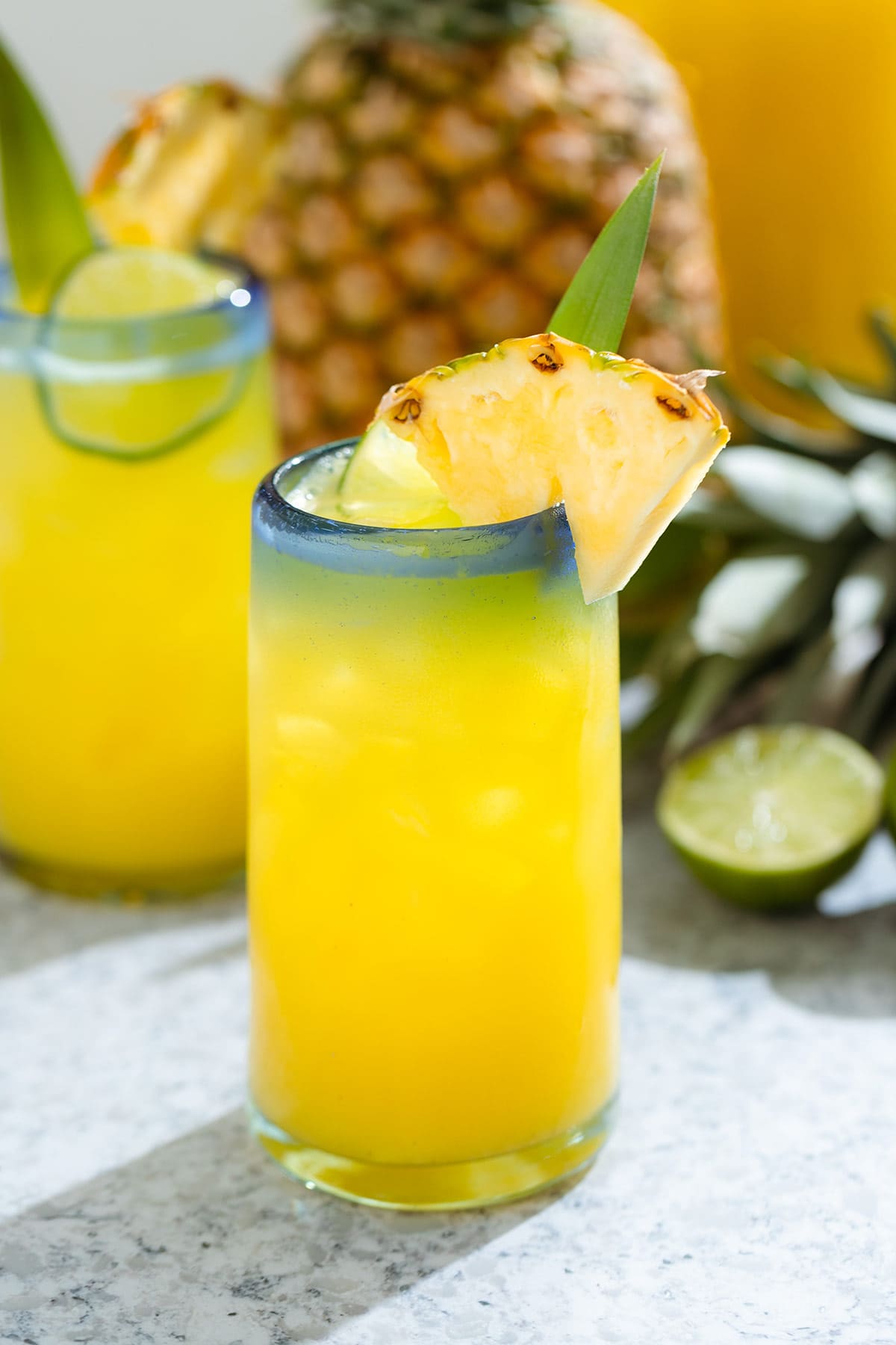 Pineapple agua fresca in a tall glass with a blue rim garnished with pineapple, lime, and a pineapple leaf.