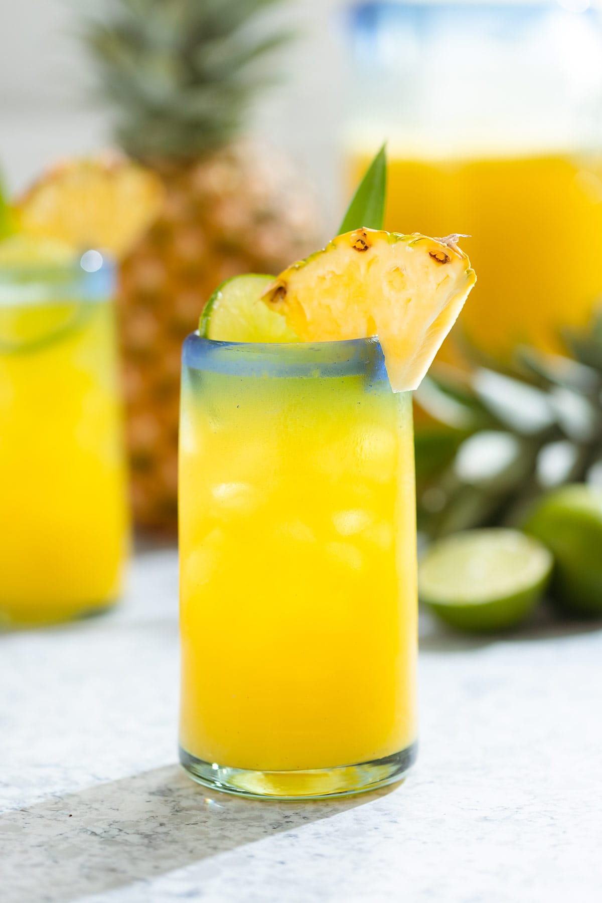Pineapple agua fresca in a tall glass with a blue rim garnished with pineapple, lime, and a pineapple leaf.