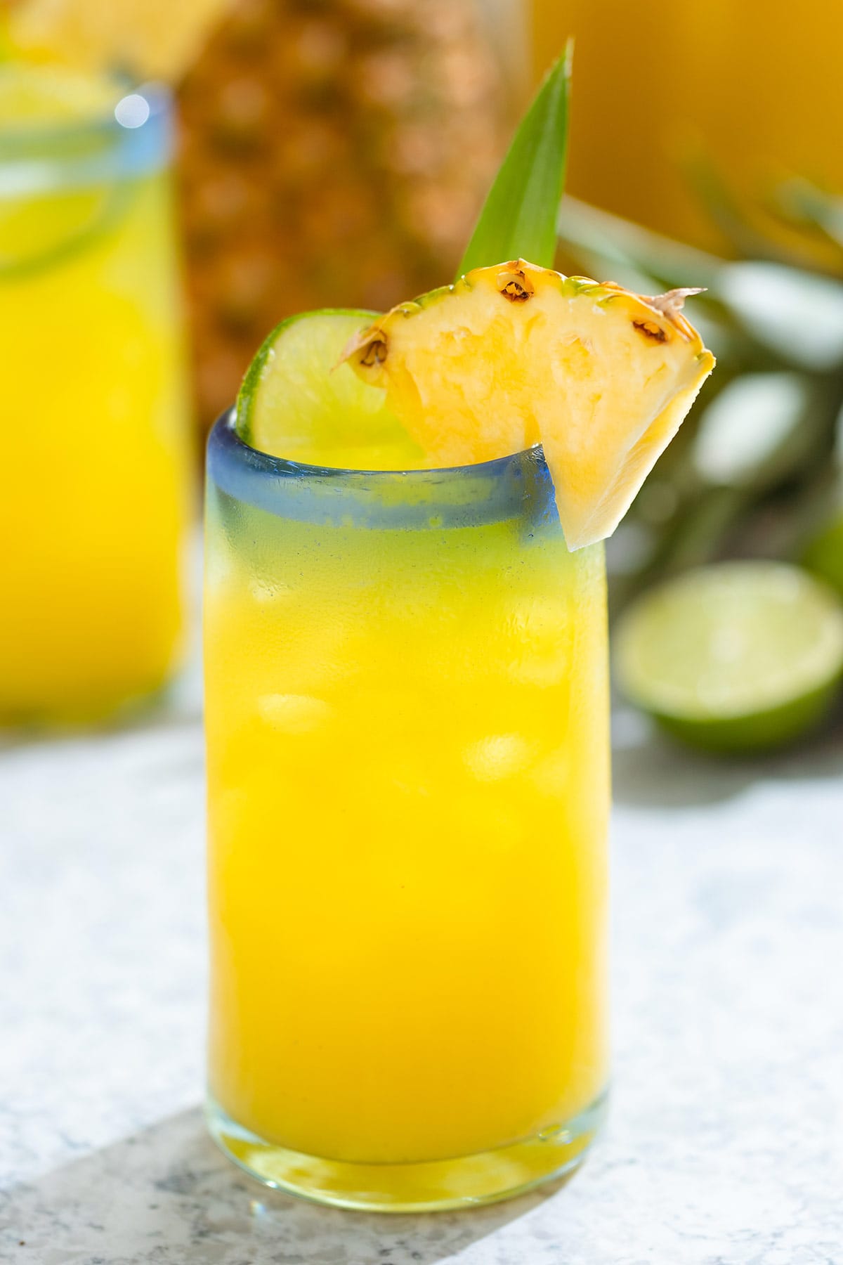 Pineapple agua fresca in a tall glass with a blue rim garnished with pineapple, lime, and a pineapple leaf.