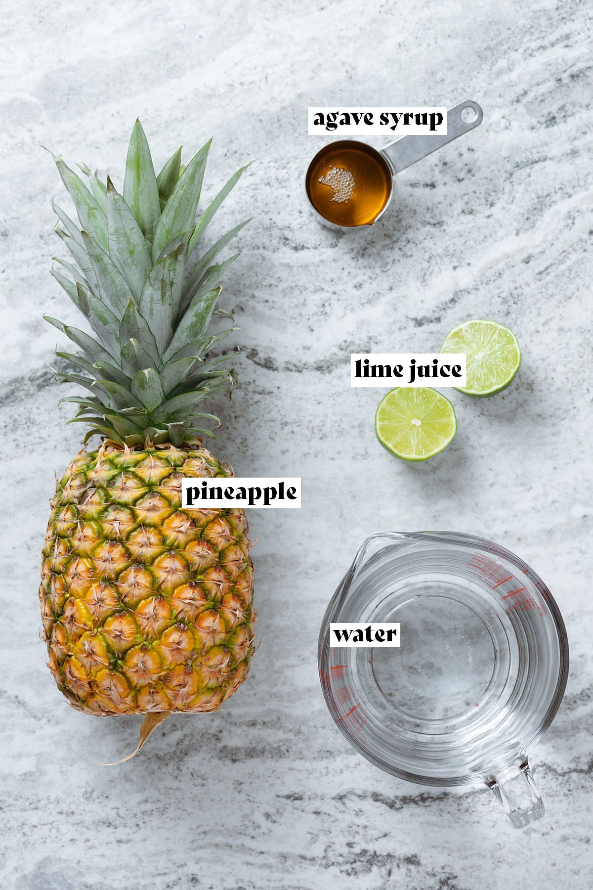 Pineapple, lime, water in a measuring cup, and agave syrup on a grey stone background.