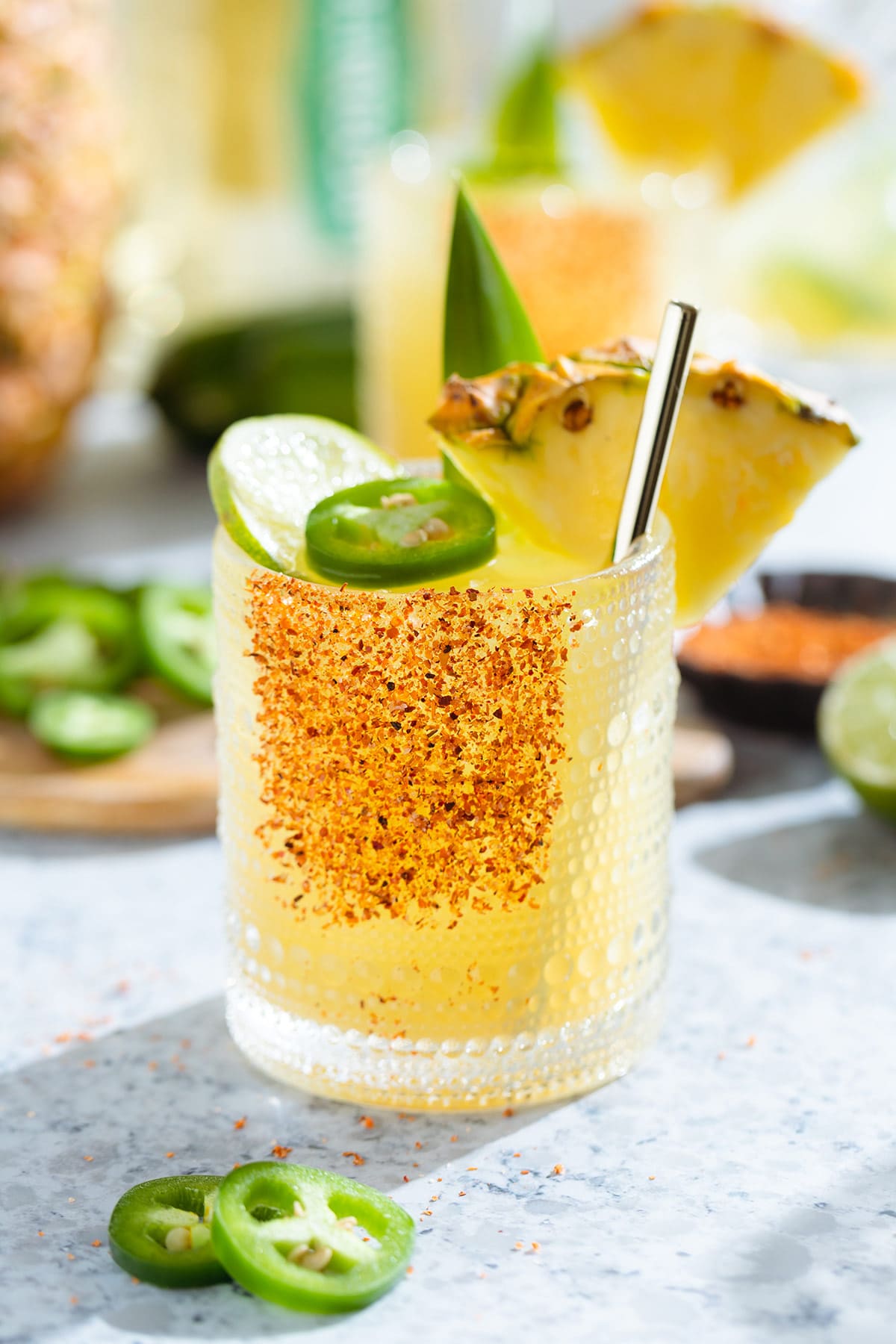 Bright yellow margarita in a short glass garnished with tajin, a wedge of pineapple, a slice of lime, and a slice of jalapeno with more fruits and drinks in the backround.