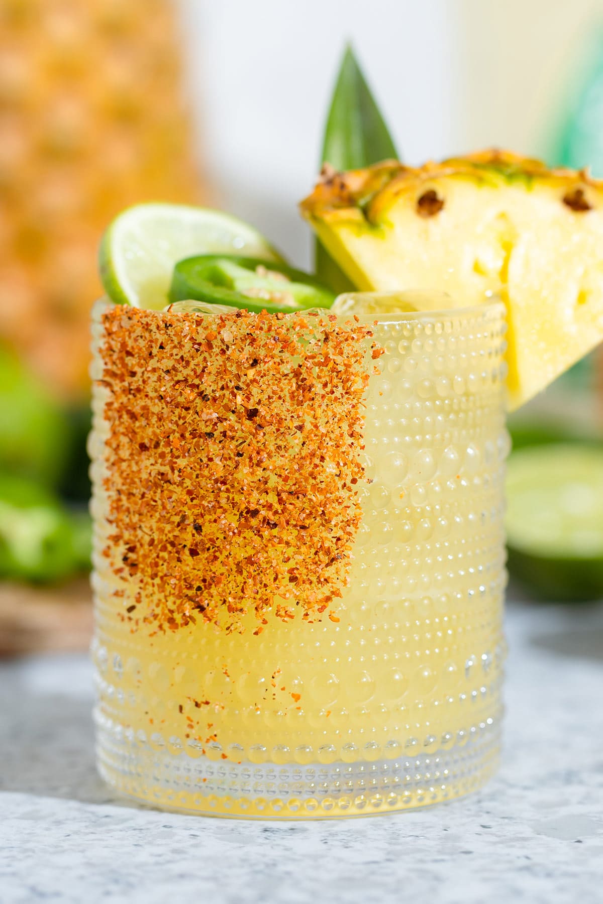 Bright yellow margarita in a short glass garnished with tajin, a wedge of pineapple, a slice of lime, and a slice of jalapeno.
