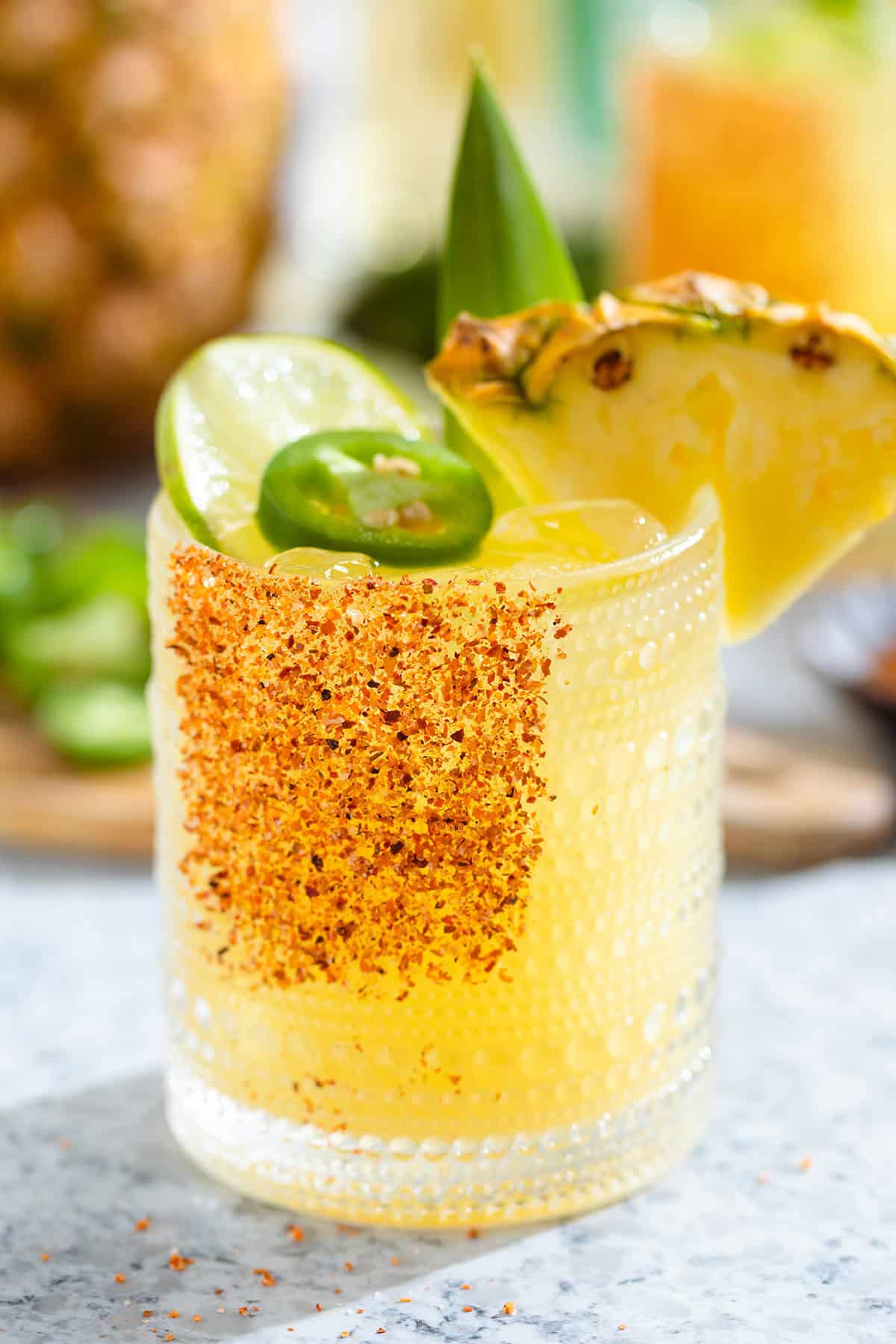 Bright yellow margarita in a short glass garnished with tajin, a wedge of pineapple, a slice of lime, and a slice of jalapeno with more fruits and drinks in the backround.