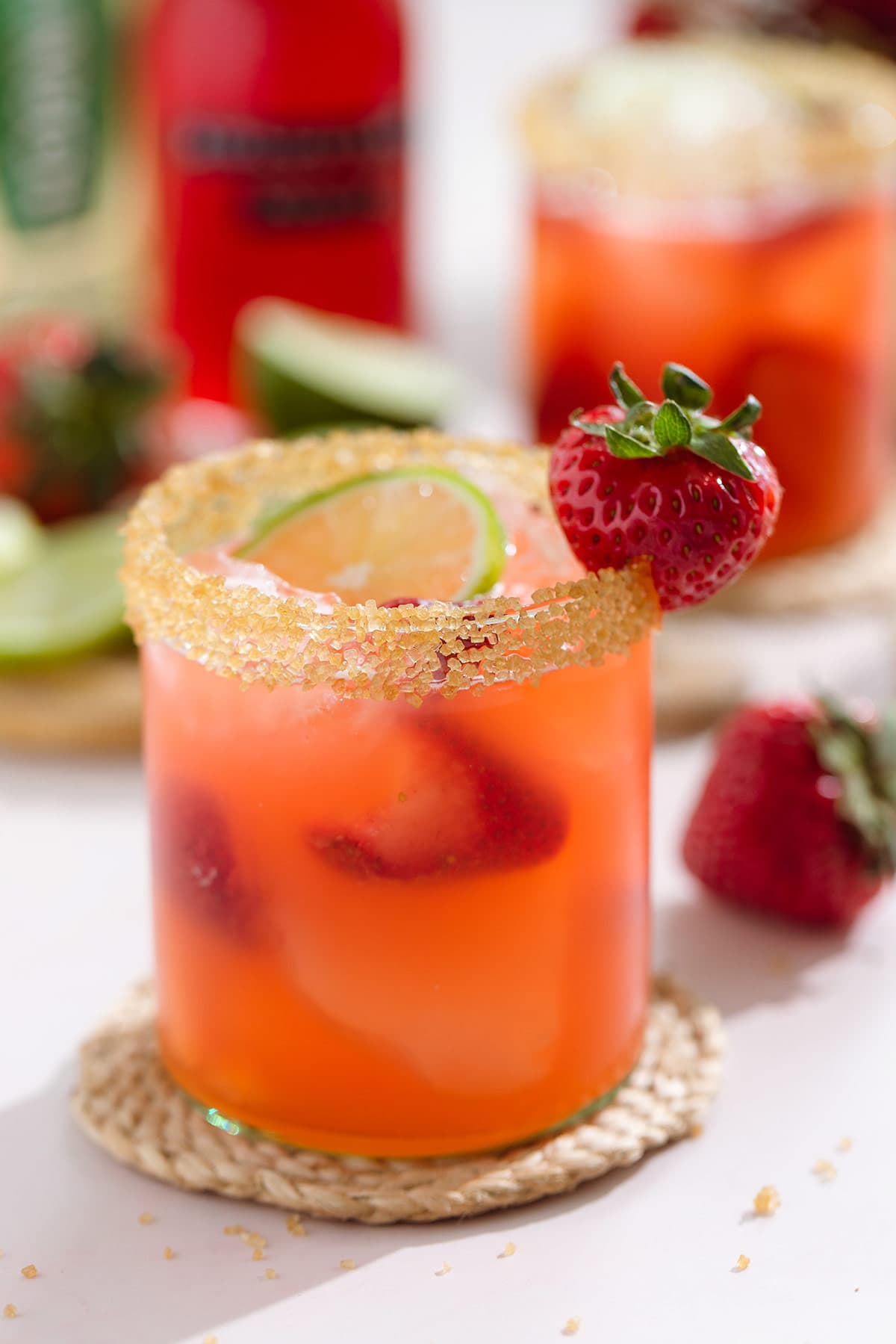 Light red margarita in a short glass garnished with coarse sugar on the rim and a strawberry on a woven coaster.