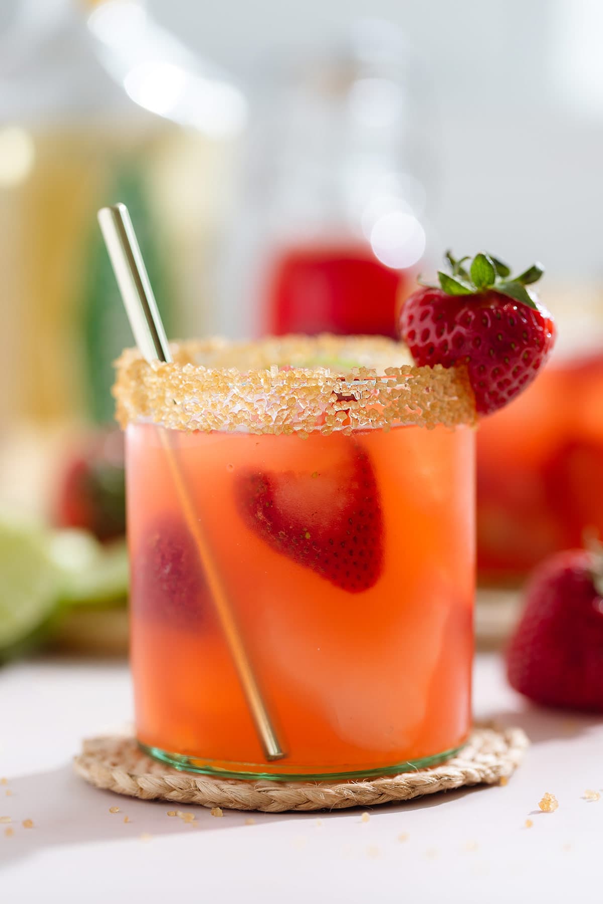 Light red margarita in a short glass garnished with coarse sugar on the rim and a strawberry with a gold straw.
