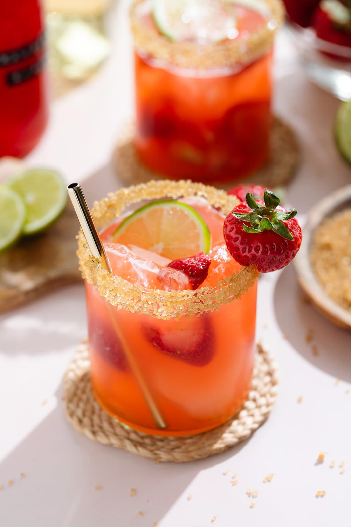 Light red margarita in a short glass garnished with coarse sugar on the rim and a strawberry with a gold straw with another margarita in the backround.