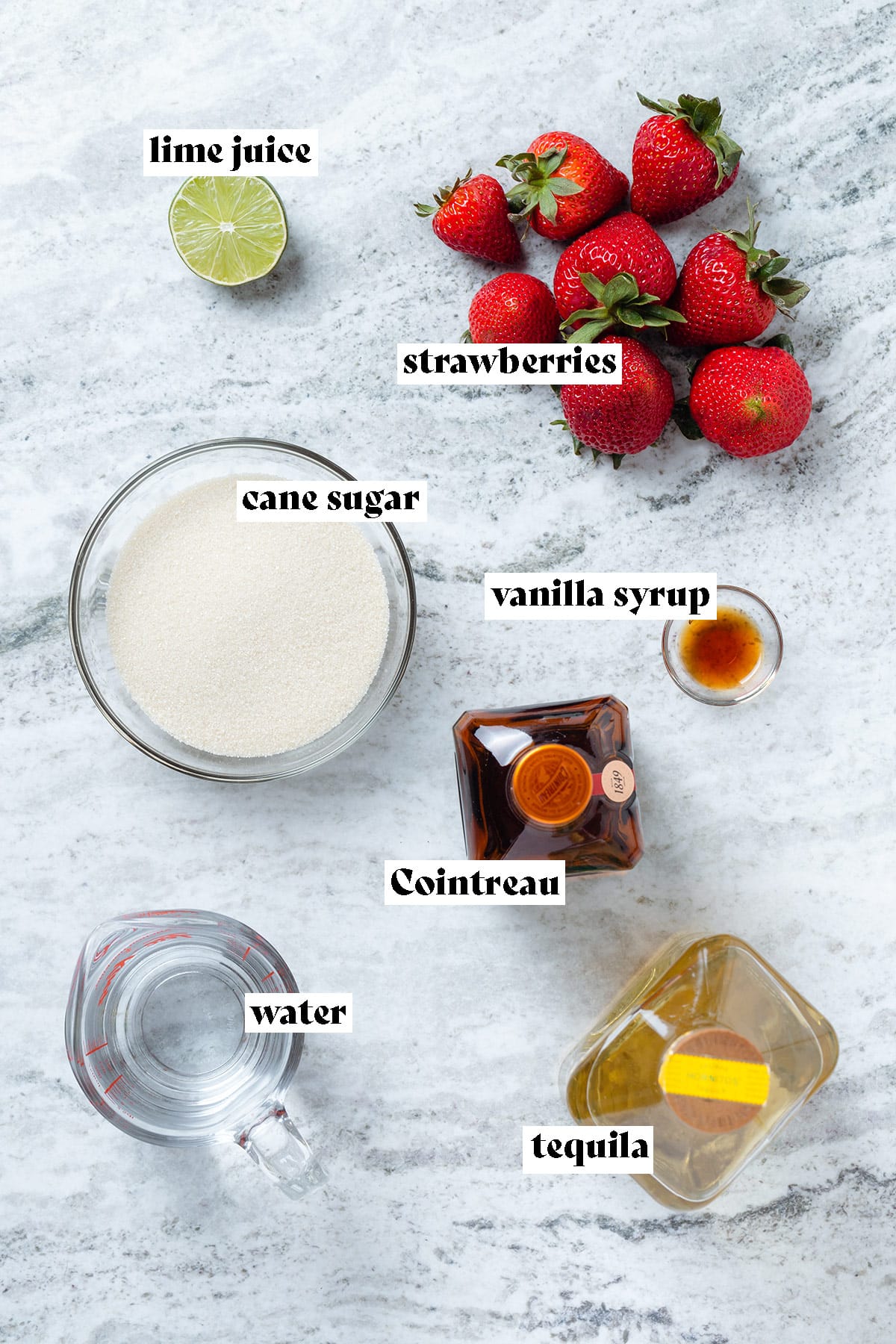 Ingredients for strawberry margarita like tequila, strawberries, and lime juice laid out on a grey stone background.