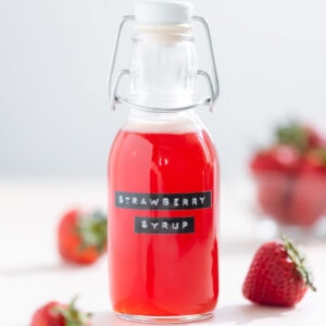 Bright red strawberry syrup in a glass jar with a label that says strawberry syrup on it, on a white background.