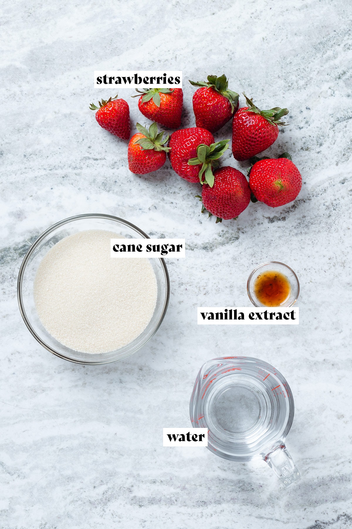 Strawberries, cane sugar, agave, and water in a glass measuring cup laid out on a grey stone background.