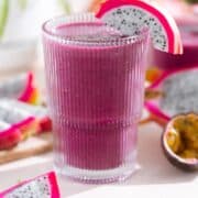 Bright purple smoothie in a tall glass garnished with white dragon fruit with more dragon fruit and passion fruit around the glass.