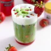 A tall glass with a matcha latte with a layer of frothy milk on the top and a layer of strawberry syrup on the bottom of the glass garnished with more chopped strawberries on top.