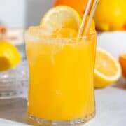 A short glass with bright orange citrus cocktail on a grey background with more lemons and oranges in the background.