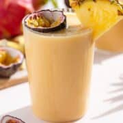 Light orange smoothie in a tall glass garnished with a slice of pineapple and half of a passion fruit.