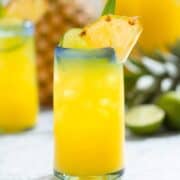 Pineapple agua fresca in a tall glass with a blue rim garnished with pineapple, lime, and a pineapple leaf.