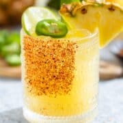 Bright yellow margarita in a short glass garnished with tajin, a wedge of pineapple, a slice of lime, and a slice of jalapeno with more fruits and drinks in the backround.