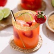 Light red margarita in a short glass garnished with coarse sugar on the rim and a strawberry with a gold straw with another margarita in the backround.