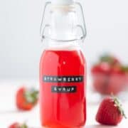 Bright red strawberry syrup in a glass jar with a label that says strawberry syrup on it, on a white background.