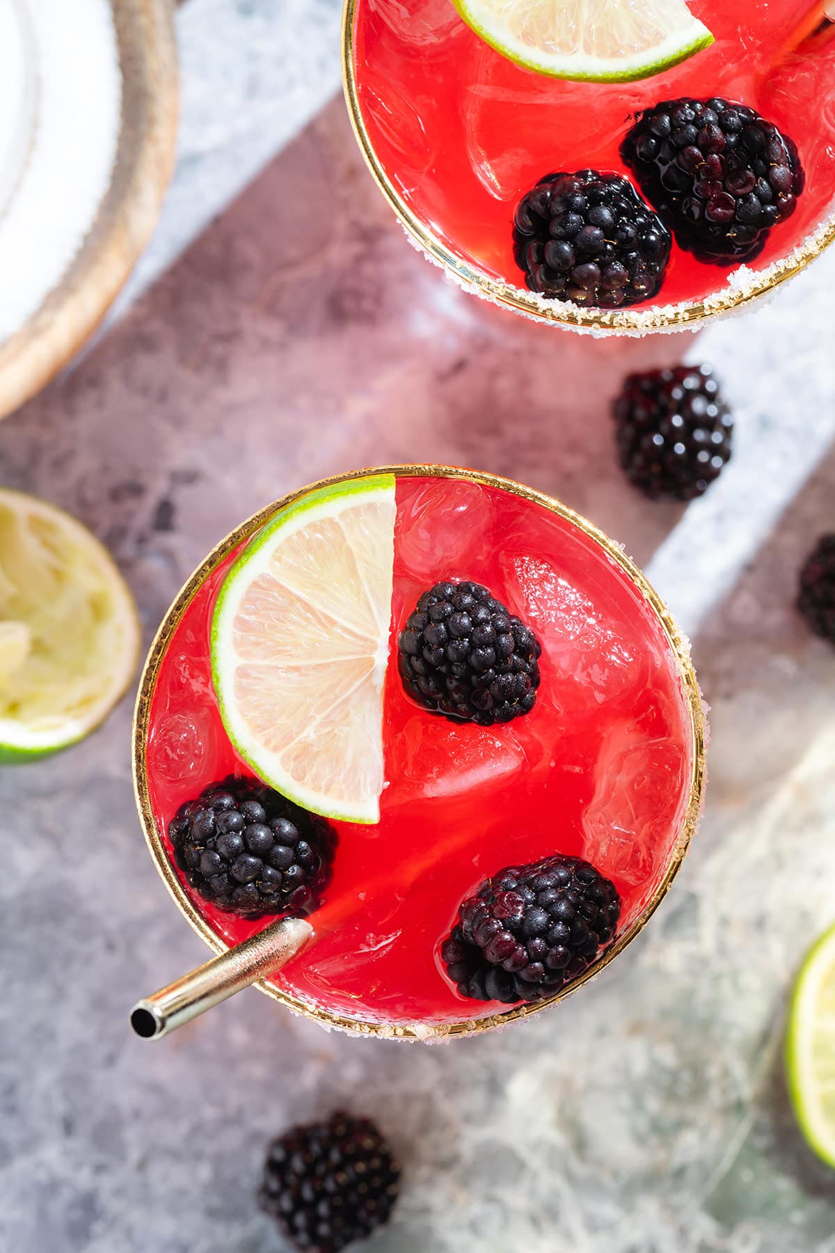 Bright purple red blackberry margarita in a glass with a gold rim garnished with blackberries and lime slices and salt on the rim shot from above with more blackberries around the glass.