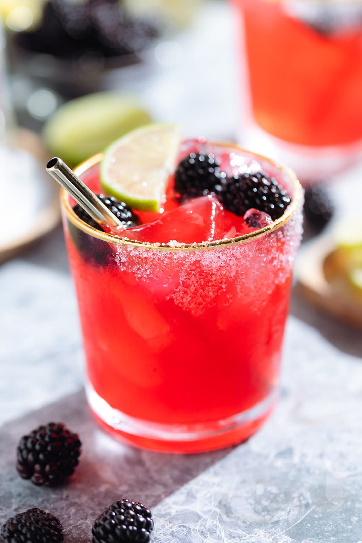 Bright purple red blackberry margarita in a glass with a gold rim garnished with blackberries and lime slices and salt on the rim on a grey stone background.
