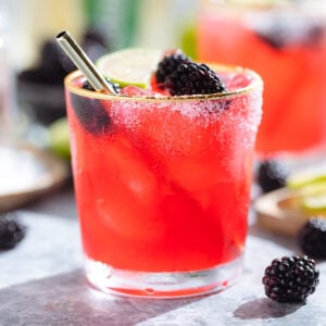 Bright purple red blackberry margarita in a glass with a gold rim garnished with blackberries and lime slices and salt on the rim.