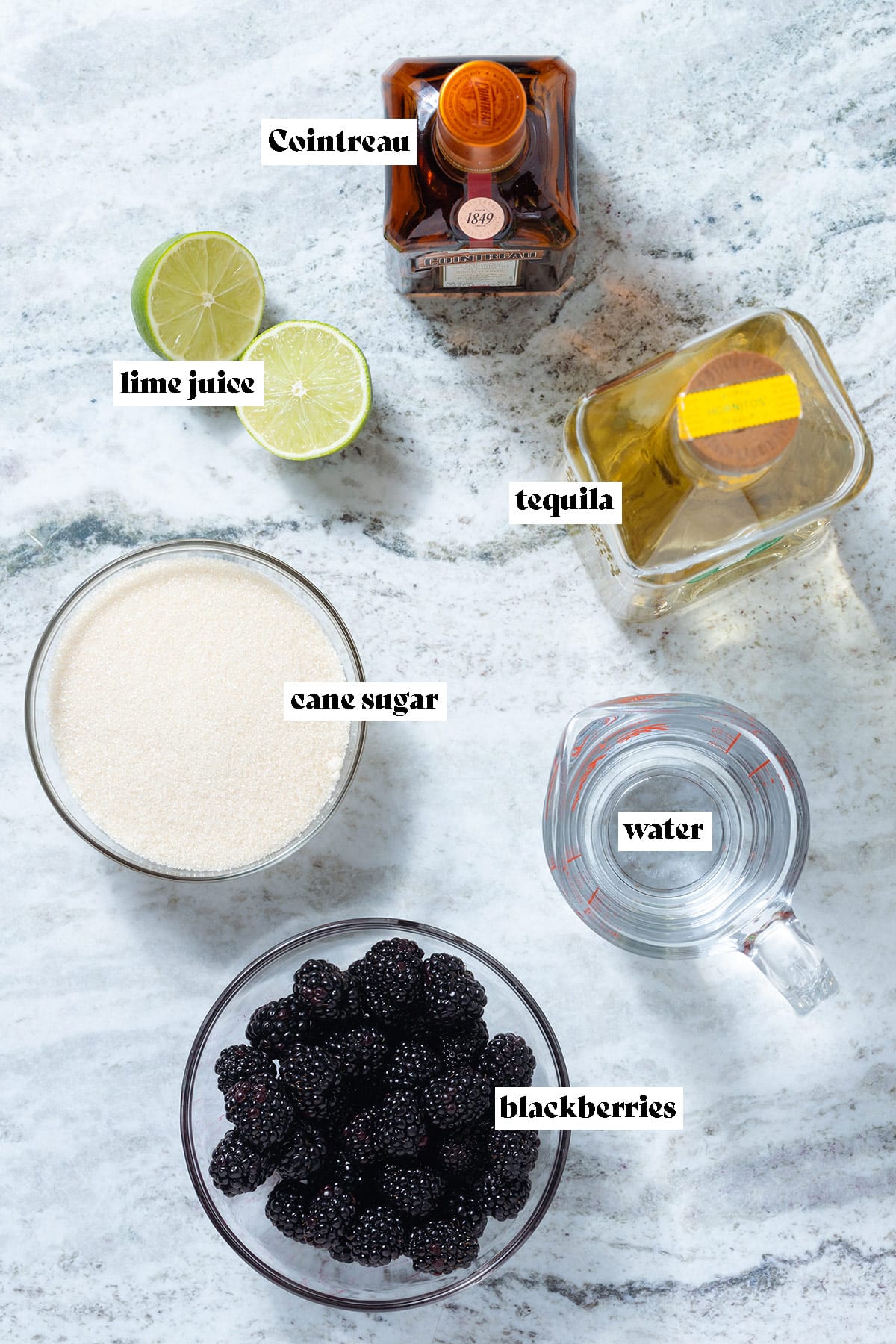 Blackberries, tequila, cane sugar, and other ingredients laid out on a grey stone background.
