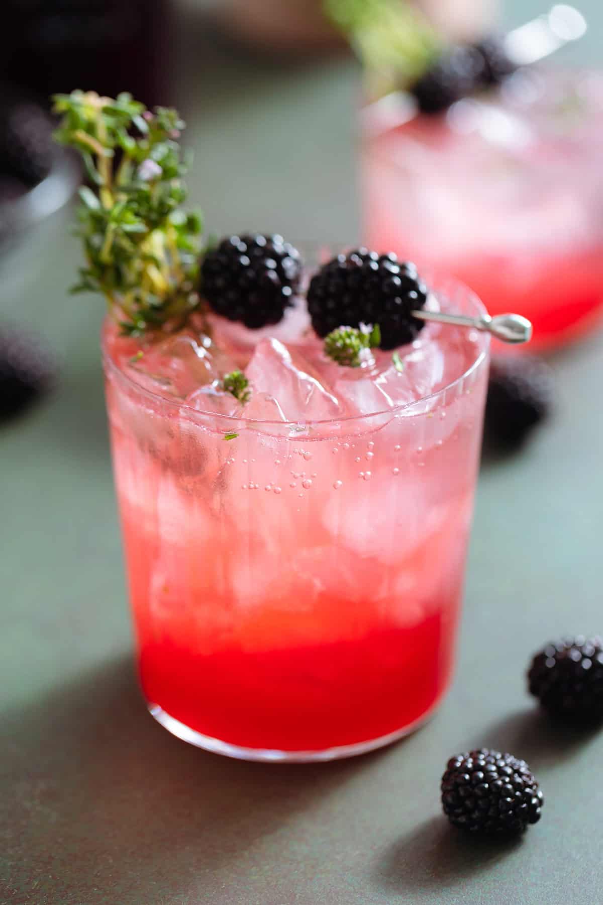 Bright red blackberry mocktail with seltzer on top creating an ombre effect in a short glass garnished with blackberries and fresh thyme.