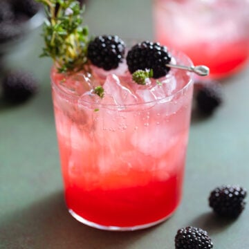 Bright red blackberry mocktail with seltzer on top creating an ombre effect in a short glass garnished with blackberries and fresh thyme.