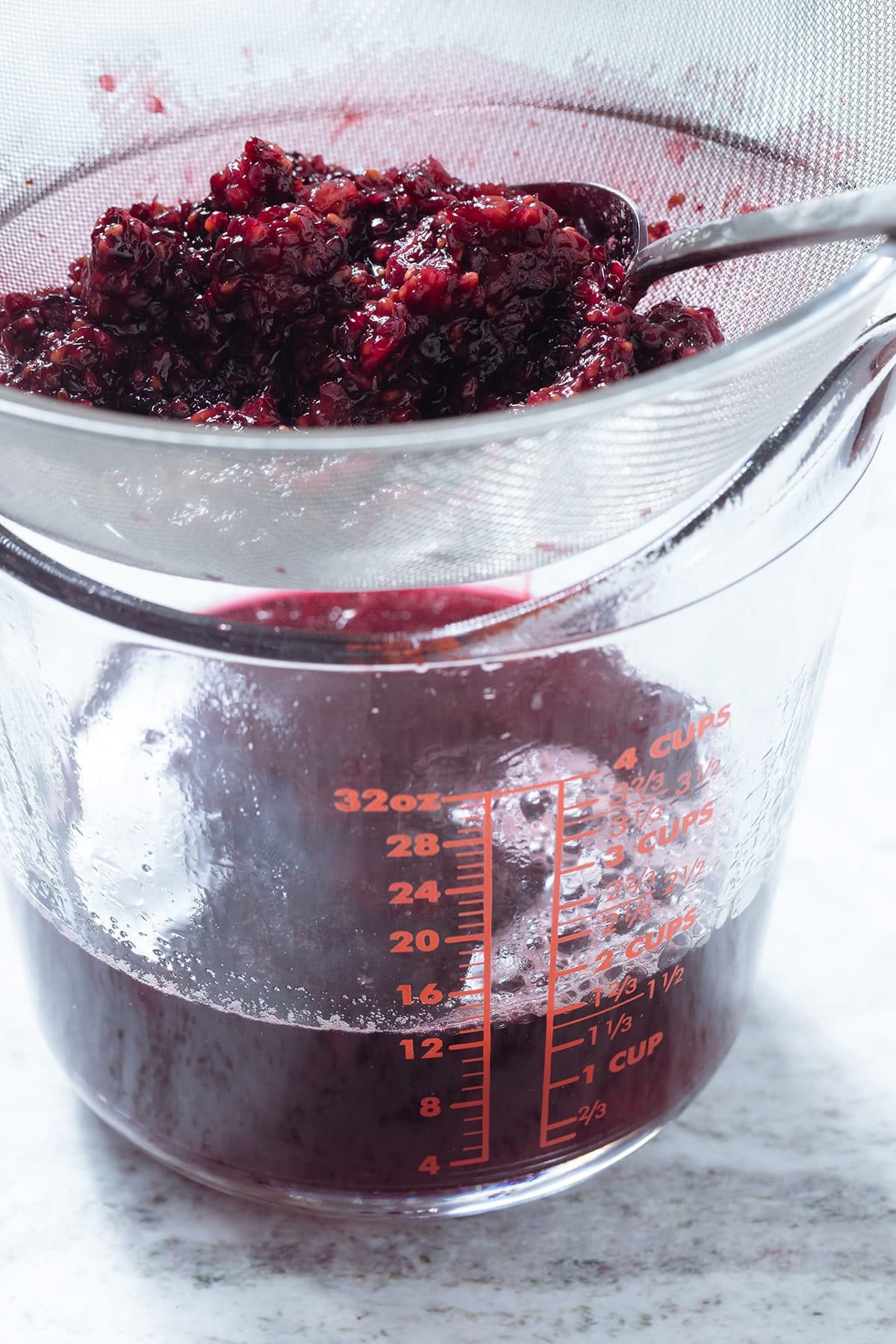 Blackberry syrup straining through a fine mesh strainer into a tall glass measuring cup.
