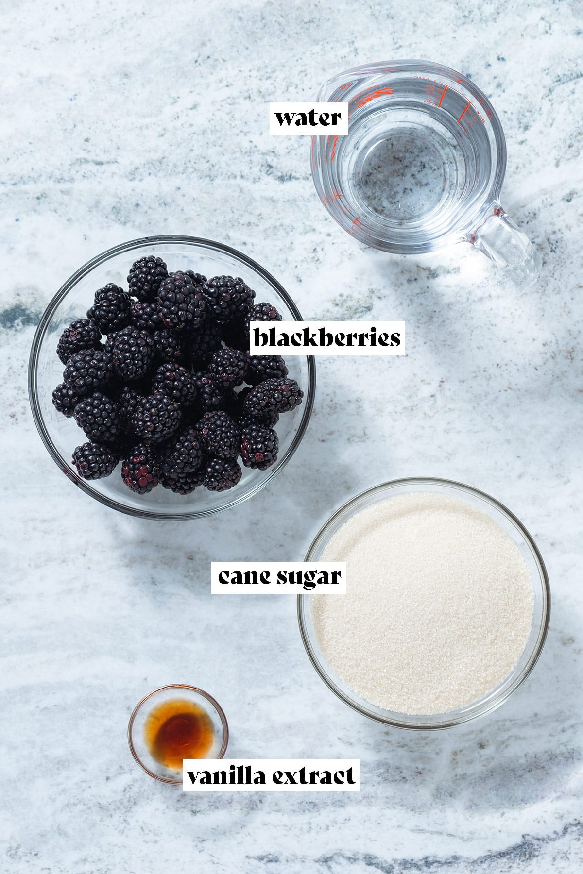 Blackberries, cane sugar, water, and vanilla extract laid out on a grey stone background.