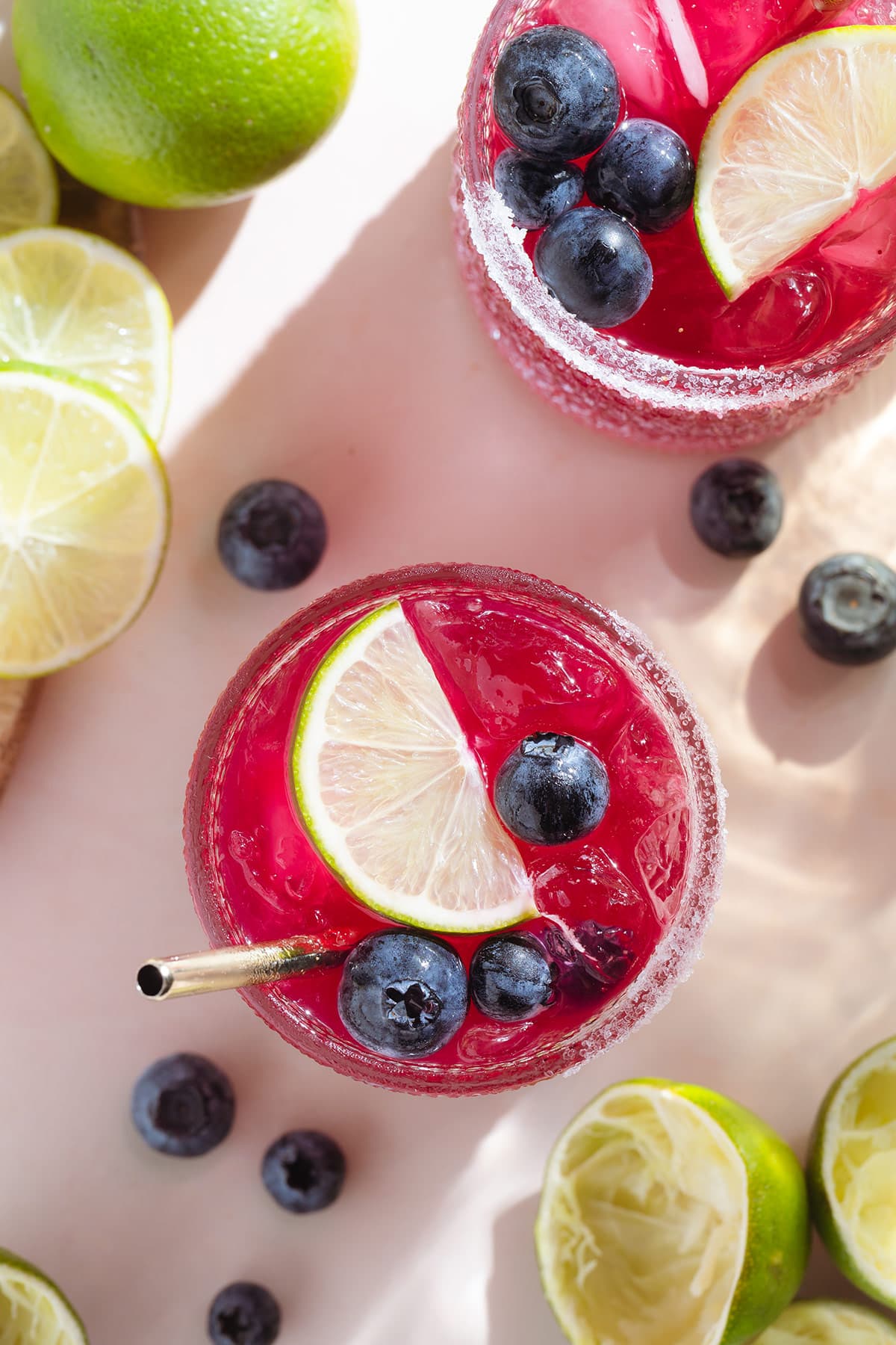 Bright pink purple margarita in a short glass garnished with blueberries and a lime slice with salt on the rim and a gold straw shot from above.
