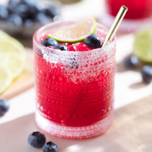 Bright pink purple margarita in a short glass garnished with blueberries and a lime slice with salt on the rim and a gold straw.