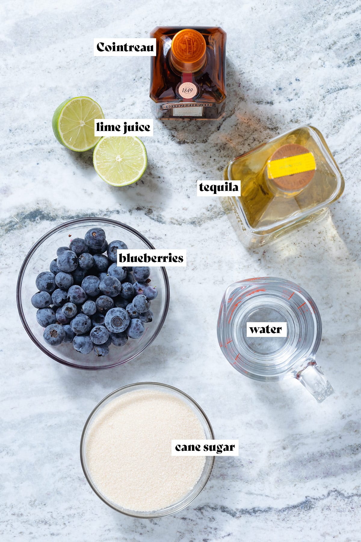 Blueberries, tequila, cointreau, limes, and other ingredients on a grey stone background.