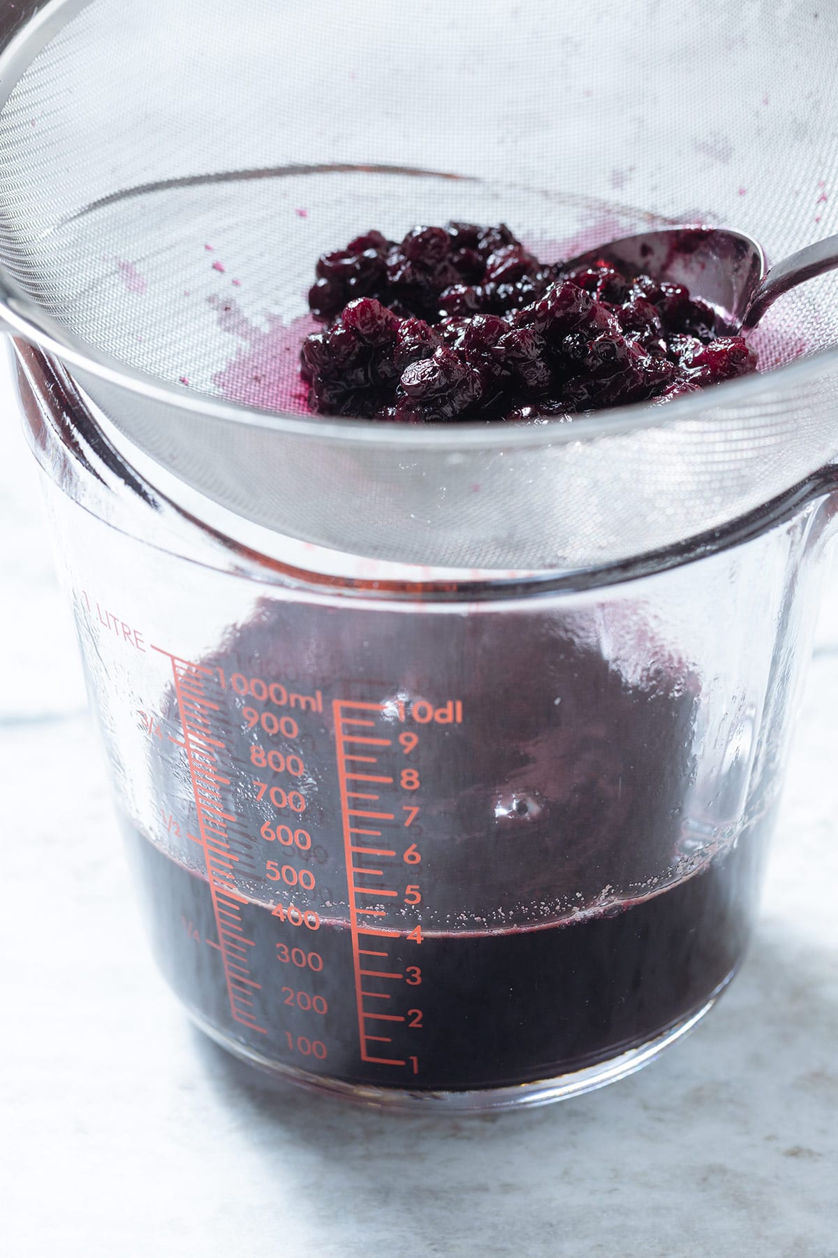 Blueberry syrup being strained through a fine mesh strainer into a tall glass jar.