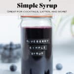 A glass jar with dark purple blueberry syrup with fresh blueberries around the jar and a green plant in the background.