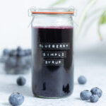 A glass jar with dark purple blueberry syrup with fresh blueberries around the jar and a green plant in the background.