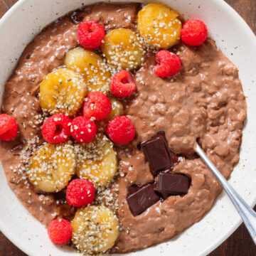 Chocolate oatmeal in a beige bowl topped with fresh raspberries, caramelized banana slices and chocolate squares.