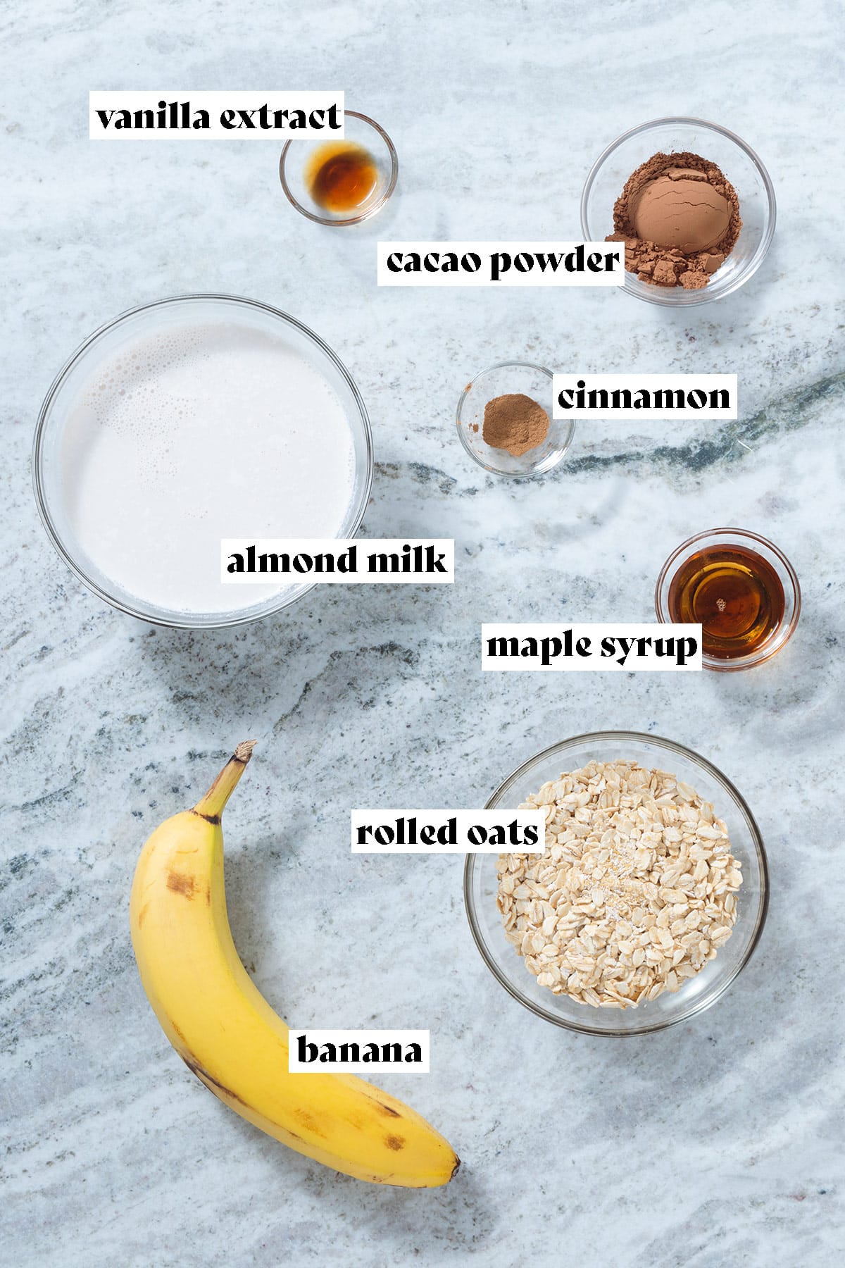 Rolled oats, almond milk, banana, and other ingredients laid out on a grey stone background.