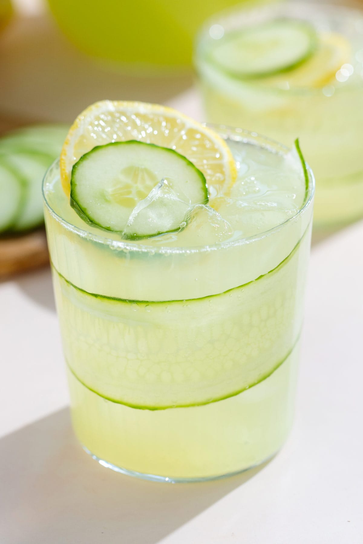 A short glass with light green cucumber lemonade over ice garnished with a slice of lemon and cucumber with a thin strip of cucumber around the inside wall of the glass.