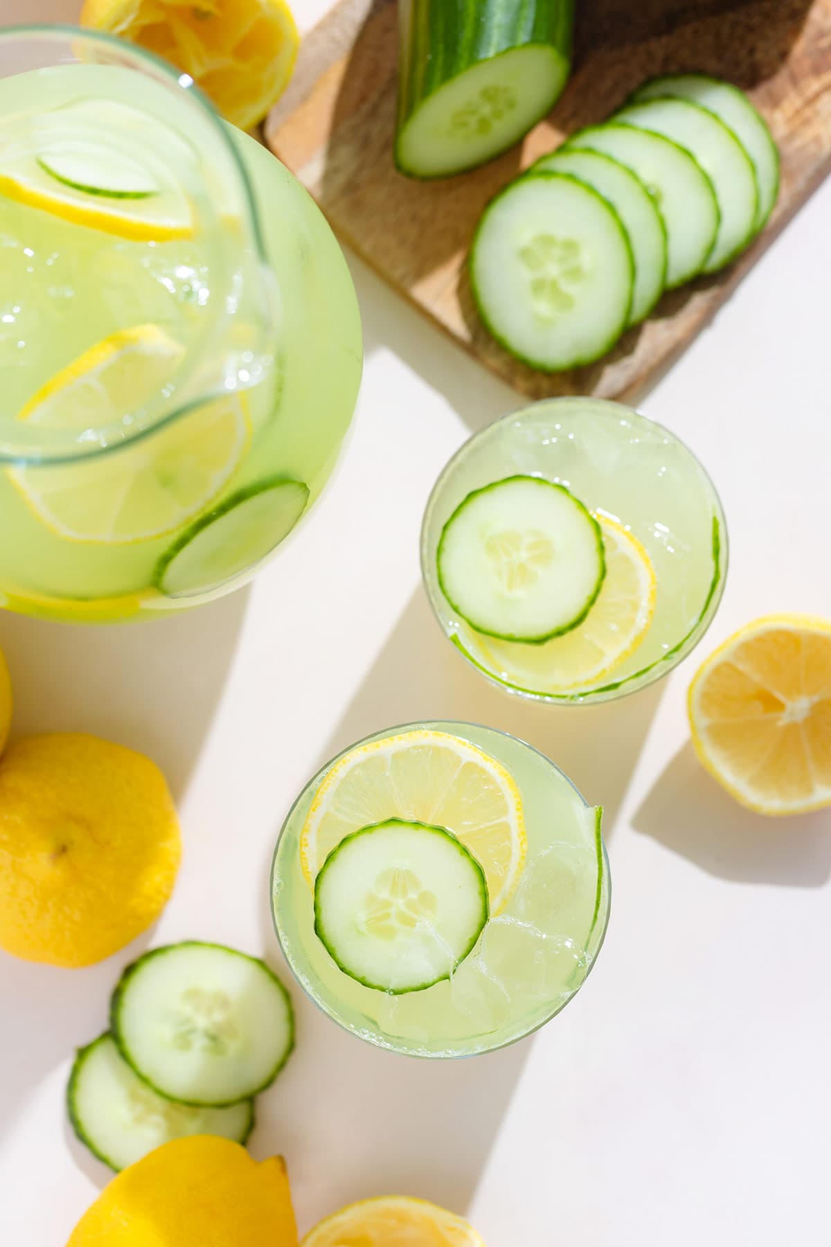 Two glasses glass with light green cucumber lemonade over ice garnished with a slice of lemon and cucumber with more lemonade and cucumber around.