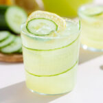 A short glass with light green cucumber lemonade over ice garnished with a slice of lemon and cucumber with more lemonade in the background.