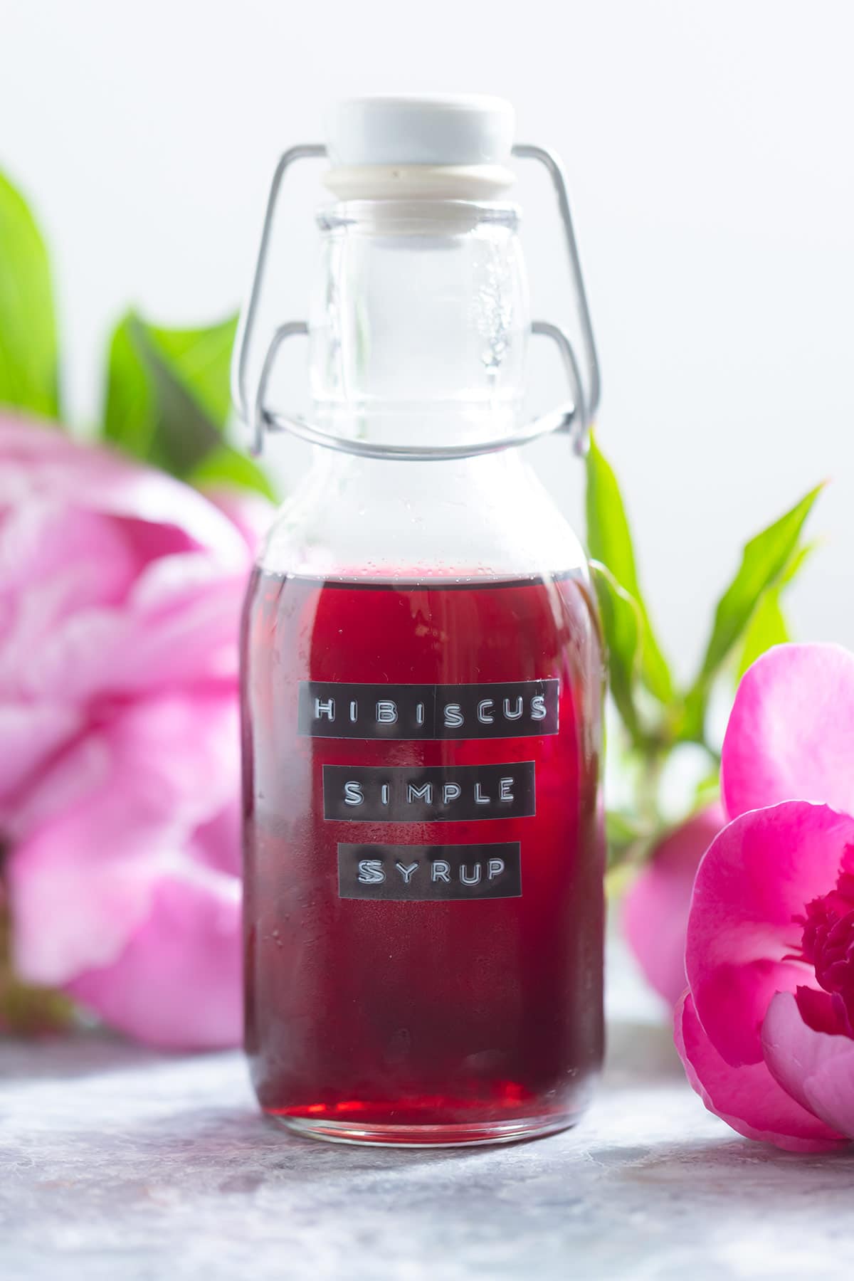 A small glass bottle with bright red hibiscus syrup with peonies in the background.