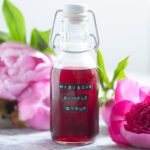A small glass bottle with bright red hibiscus syrup with peonies in the background.