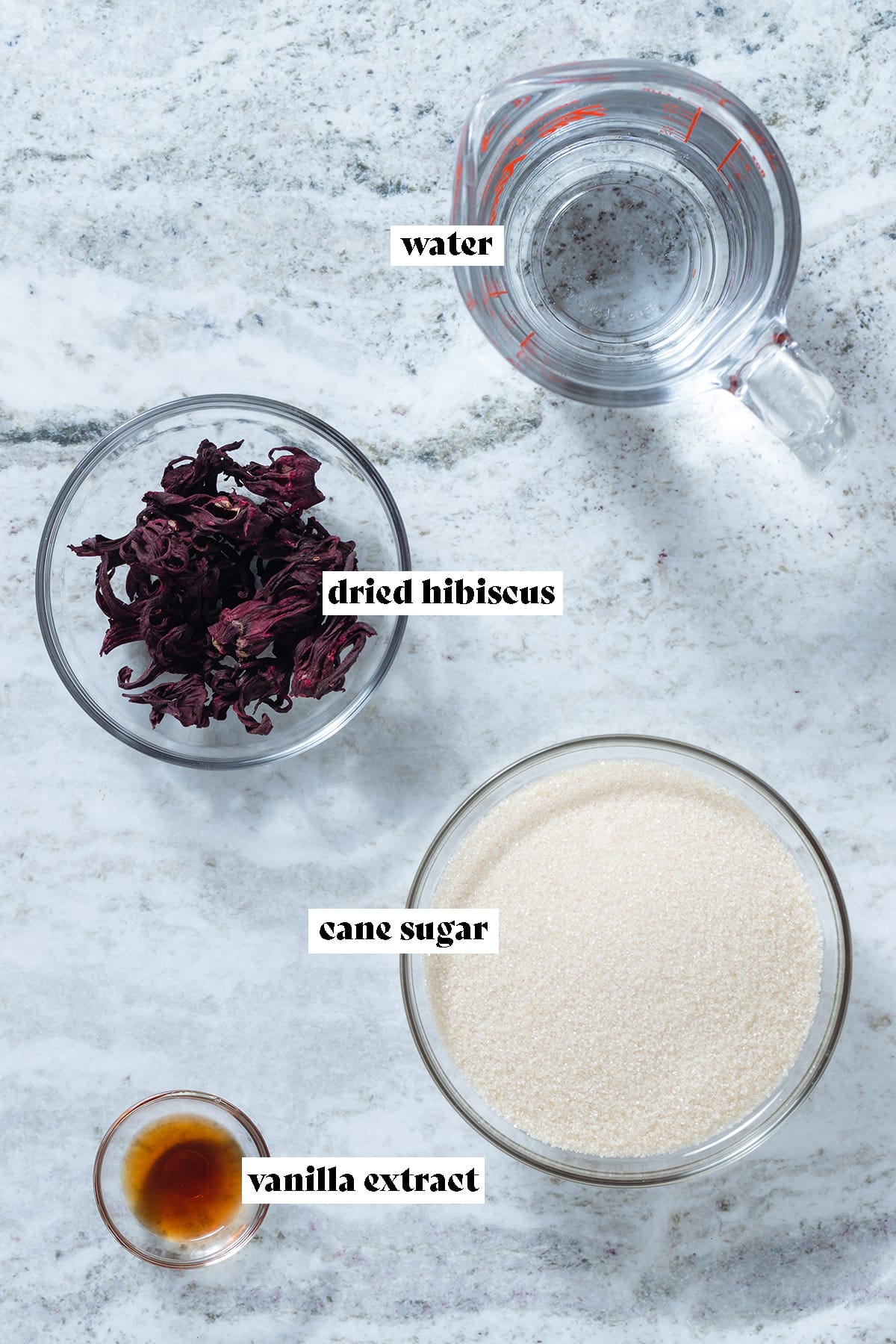 Dried hibiscus flowers, water, cane sugar, and vanilla extract laid out on a grey stone background.